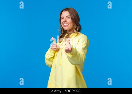 Hey you. Cheeky and carefree cute modern european girl in yellow hoodie, wink and make finger pistols gesture at camera to say h Stock Photo