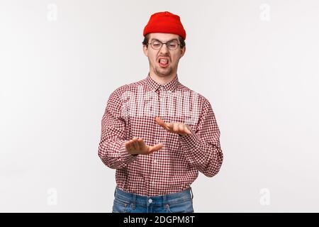 Portrait funny relucant young man in red beanie and glasses, shaking hands in rejection, stick tongue and grimacing disgusted, e Stock Photo