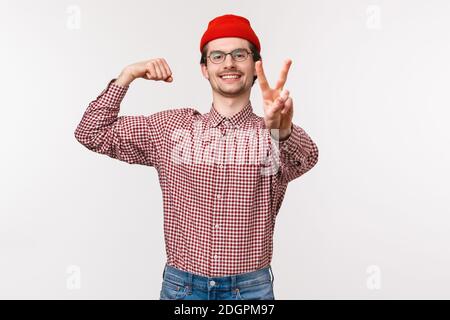 Waist-up portrait young carefree smiling man in glasses and beanie, showing peace gesture and flex biceps with satisfied express Stock Photo