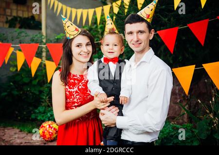 Theme children birthday party. Family father and mother holding son of one year on the background of greenery and festive decor, Stock Photo