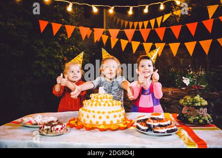 Children's birthday party. Three cheerful children girls at the table eating cake with their hands and smearing their face. Fun Stock Photo