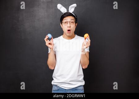 Holidays, party and Easter concept. Amazed young asian guy in rabbit ears holding painted eggs as learning girlfriend traditions Stock Photo