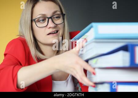 Businesswoman with eyeglasses holding large pile of folders with documents in office Stock Photo