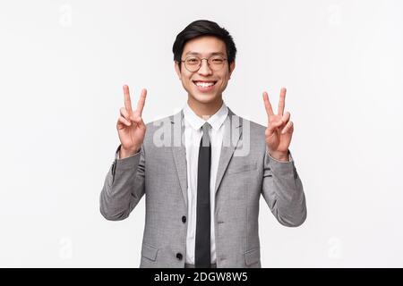 Waist-up portrait of optimistic cheerful and handsome asian man in grey suit, glasses showing peace signs, smiling kawaii, takin Stock Photo
