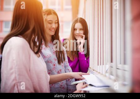 Three cheerful high school students doing homework together while standing near the window of the school. Stock Photo