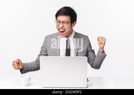 Portrait of successful pleased young asian man in grey suit, wireless headphones, close eyes saying yes, shouting from pleasure Stock Photo
