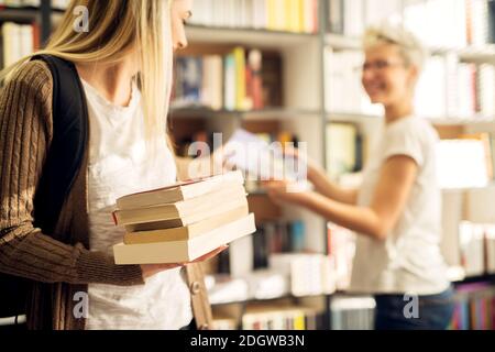 A young cheerful female student is taking the book from a female dedicated joyful librarian and stacking them in other hand. Stock Photo