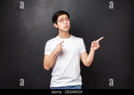 Portrait of skeptical and displeased young picky asian guy looking doubtful at product, being unsure in quality, pointing finger Stock Photo