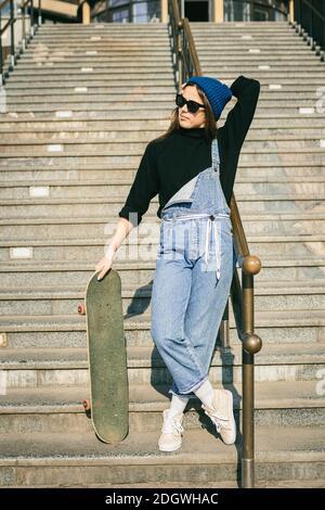 Young caucasian woman posing on street with skateboard in hands. Teenager girl in blue jeans extreme sports in an urban environm Stock Photo