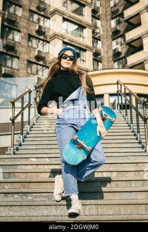 Stylishly dressed woman in blue denim jumpsuit posing with skateboard. Street photo. Portrait of girl holding skateboard. Lifest