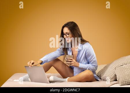 Attractive young woman in blue shirt, wearing glasses, sitting on bed, in front of her laptop, drinking a cup of tea Stock Photo