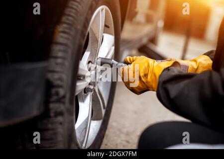 An experienced hard working mechanic in orange gloves is using an electric wrench to tighten up the screws in a car wheel. Stock Photo