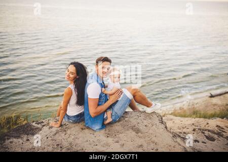 Theme family vacation with small child on the nature and sea. Mom, Dad and daughter of one year are sitting in embrace, girls in Stock Photo