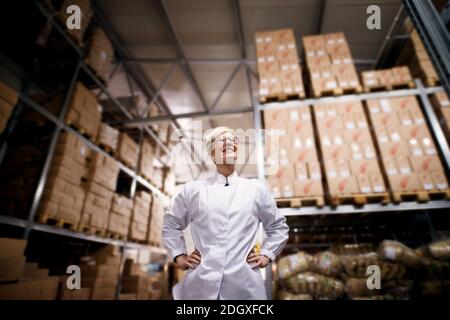 Young beautiful proud female worker is holding her arms on her hips and smiling in a storage room of a factory. Stock Photo