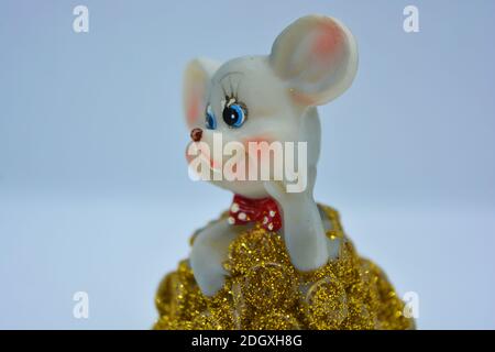 A figurine of a cute gray mouse peeking out of a mountain of yellow coins is located on a white fabric background. Stock Photo