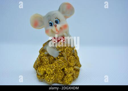A figurine of a cute gray mouse peeking out of a mountain of yellow coins is located on a white fabric background. Stock Photo