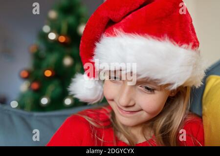 Cute blonde caucasian girl 7 years old sitting on couch near Christmas tree and writing letter to Santa Claus. Bedroom decorated with garlands. Xmas Stock Photo