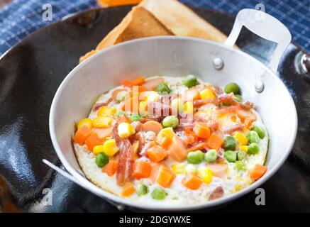 Breakfast with fried eggs in a pan with bacon and vegetables on dark plate. Stock Photo