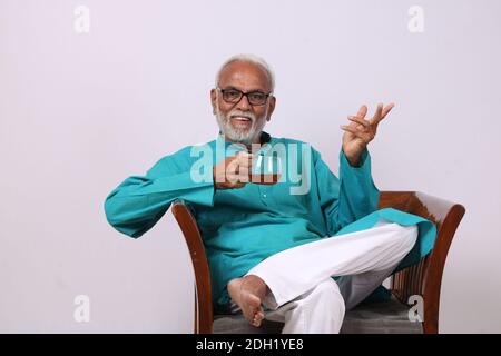 Indian old man having green tea while sitting on chair. Stock Photo