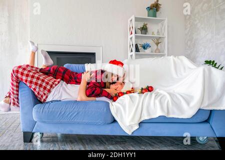 Side view of happy couple lying on couch or sofa wearing santa hats and pajamas. Young man and woman cuddling and looking at each other with tendernes Stock Photo