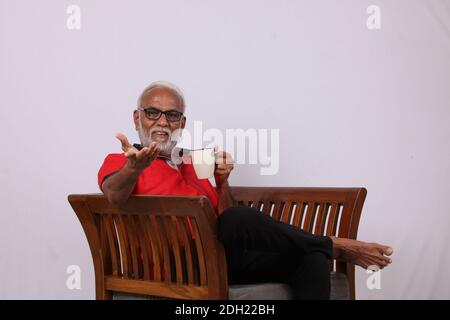 Indian old man reading magazine while sitting on chair with a coffee mug. Stock Photo