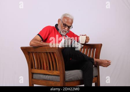 Indian old man reading magazine while sitting on chair with a coffee mug. Stock Photo