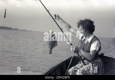 1950s, historical, out at sea, in a wooden boat, a lady wearing a patterned  skirt, top and short sleeve woollen cardigan, fishing, sitting holding a fishing  rod and holding up her catch