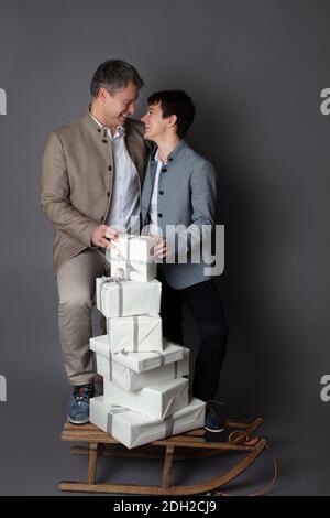 Christmas themed vertical business couple portrait dressed smart casual in blue, white, beige pants and blazers on grey background. In the foreground a wooden sleigh with a pile of white gift boxes on it. Stock Photo
