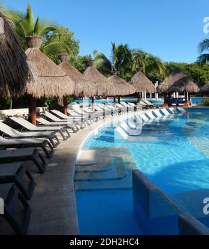 Pool sitting area with straw umbrellas, chaise longue chairs and palm trees landscape, at a Caribbean beach resort on the Riviera Maya, in Cancun, Mex Stock Photo