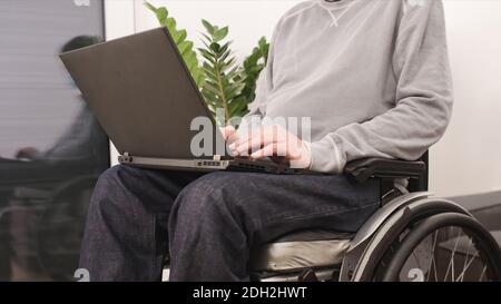 Young man in wheelchair with laptop working remotely from home, using internet and looking at screen. Stock Photo