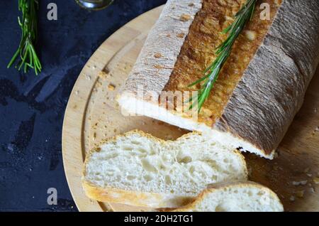 Italian ciabatta bread cut in slices on wooden chopping board with herbs, rosemary garlic and olives over dark grunge backdrop, top view.