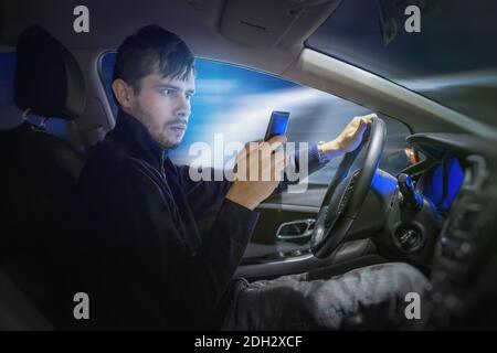 Young man is texting using smartphone and driving a car at night. Stock Photo