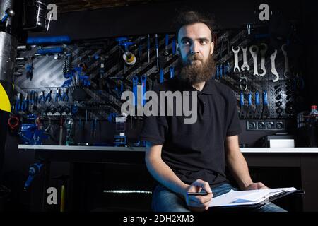 Stylish man with beard, cycle mechanic takes notes, checklist before repairing and servicing bike in bicycle shop workshop. Cauc Stock Photo