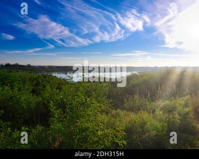 Pond Seen from Hill Surrounded by Wooded Forest and Green Foliage and Fauna with Bright Blue Sky and Wispy Clouds with Sun Rays Coming Down from Sky Stock Photo