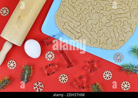 Baking Christmas cookies for dogs. Cookie cutters in shape of bones, bells, and stars and rolled out dough next to rolling pin, egg and festive decora Stock Photo