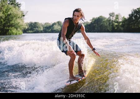Young athletic man with long hair wakesurfing on waves of river in sunny summer weather. Ttheme outdoor activities in summer. Wa