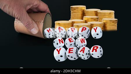 Dice spelling Happy New Year when tumbling out of a raffle cup hold by one hand  with stacks of golden coins on black background. Good luck and wealth Stock Photo