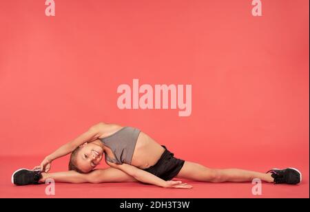 Adorable female child looking at camera and smiling while stretching and doing splits against red background Stock Photo