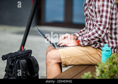 Close up of man hand typing on laptop keyboard while sitting on wooden bench in downtown. The male freelancer uses laptop and an Stock Photo