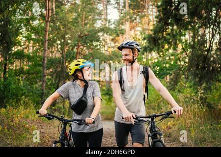 Cheerful couple pushing bicycles and walking along the forest road. Happy couple with bicycle walking through park and talking a Stock Photo