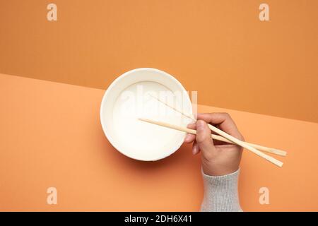 Pair of wooden chopsticks in a female hand and empty paper plate on a brown background Stock Photo