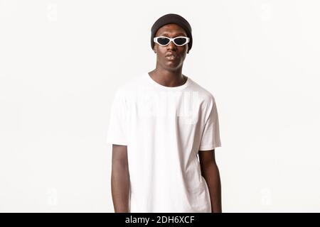 Portrait of stylish and cool african-american young guy in beanie and sunglasses, looking confident and sassy, standing over whi Stock Photo