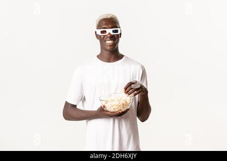 Portrait of happy attractive african-american blond guy eating popcorn and watching movie in 3d glasses, smiling cheerful, stand Stock Photo