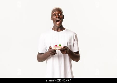 Portrait of excited, happy african-american blond guy, open mouth fascinated, holding piece of cake, standing white background Stock Photo