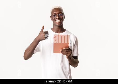 Portrait of happy good-looking african american blond male student, guy in glasses showing thumbs-up while using digital tablet, Stock Photo