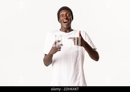 Portrait of young excited african-american guy recommending smartphone app, pointing finger at mobile phone with amazed and happ Stock Photo