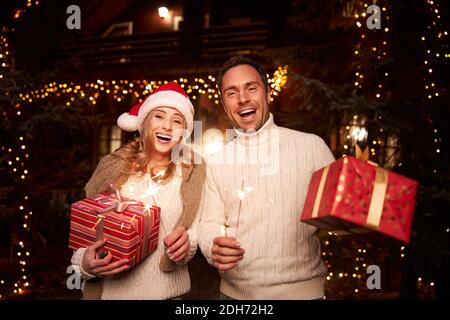 Beautiful woman and handsome man in white sweaters holding sparklers in hands near home Stock Photo
