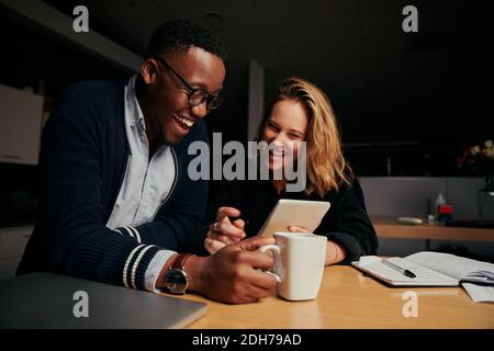 Happy businessman and businesswoman sitting together laughing while looking at digital tablet during break time at night - two colleagues smiling and Stock Photo