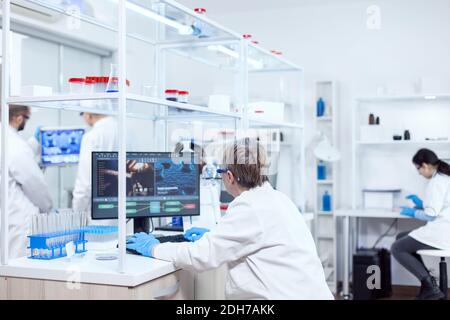 Elderly aged medical chemist working on powerful computer in modern facility. Senior scientist in pharmaceuticals laboratory doing genetic research wearing lab coat with team in the background. Stock Photo