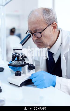 Senior scientist adjusting microscope while looking at virus and african assistant sitting at workplace. Chemist researcher in sterile lab doing experiments for medical industry using modern technology. Stock Photo